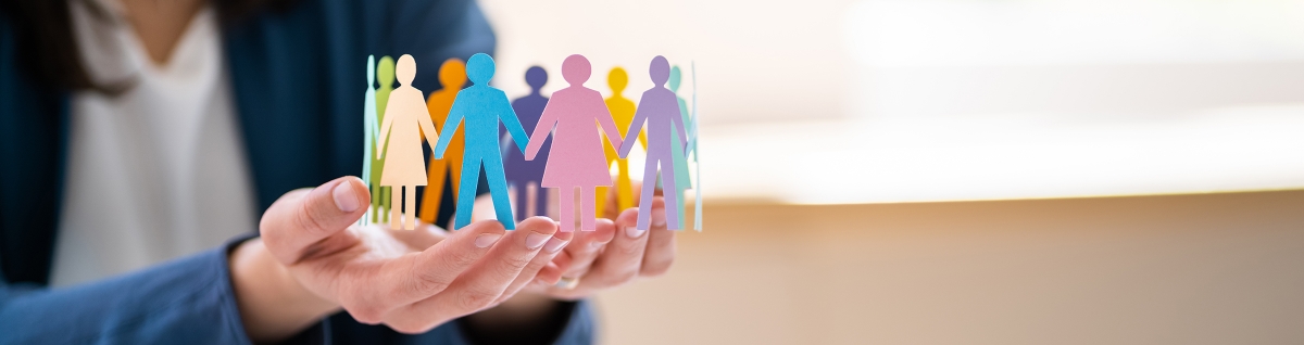 Photo: A person lovingly holds a paper craft ring in the palms of their hands. The paper craft is made of colorful paper cut out in the shape of human figures looped around in a circle as if they are holding hands.