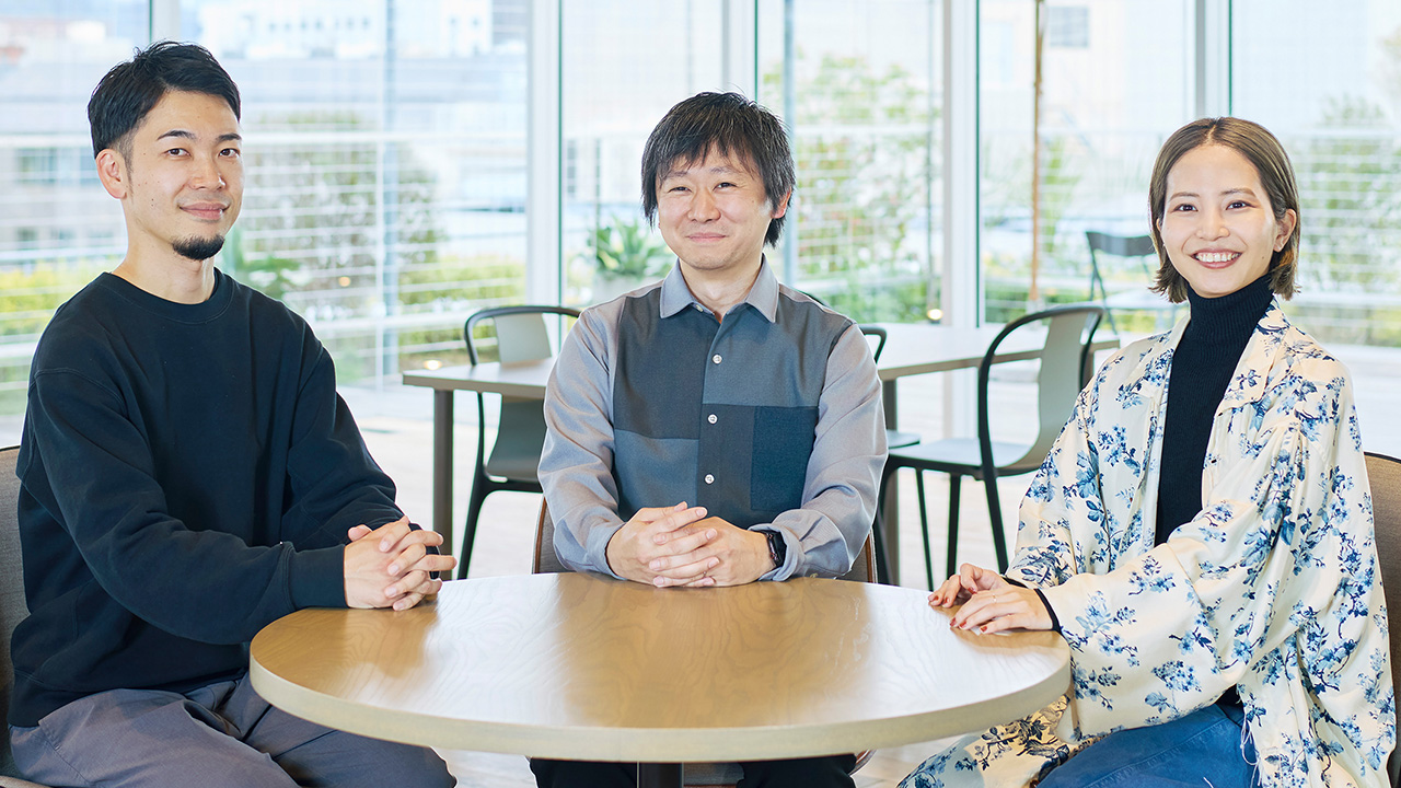 明るい室内のテーブルに3人が座っているシーン。中央に位置する男性はグレーのシャツを着用し、腕を組んでいる。左側の男性は黒のスウェットシャツを着ており、両手をテーブルに置いている。右側の女性は黒のタートルネックの上に花柄のカーディガンを羽織っている。背後には窓からの自然光が差し込み、緑が見える。