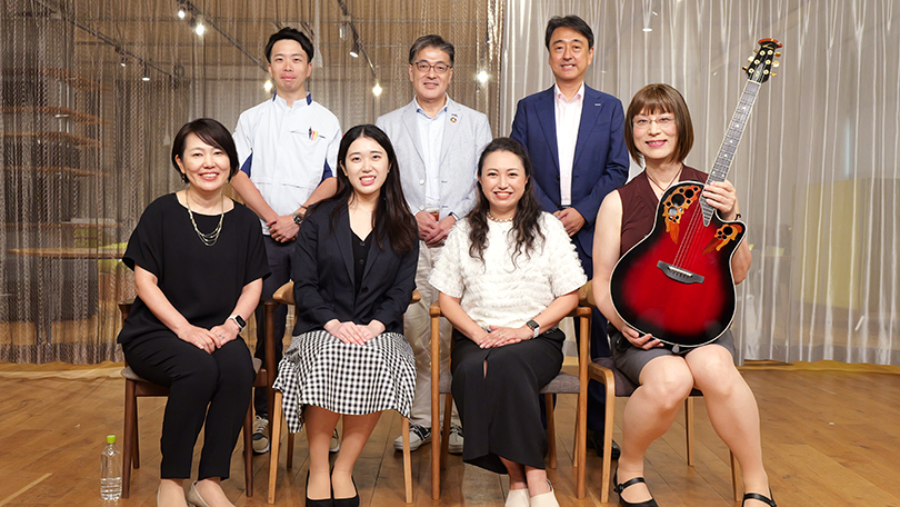 写真：笑顔で記念撮影する5人の登壇者と楠見グループCEO、玉置グループCIO。うち1人の登壇者はギターを抱えている