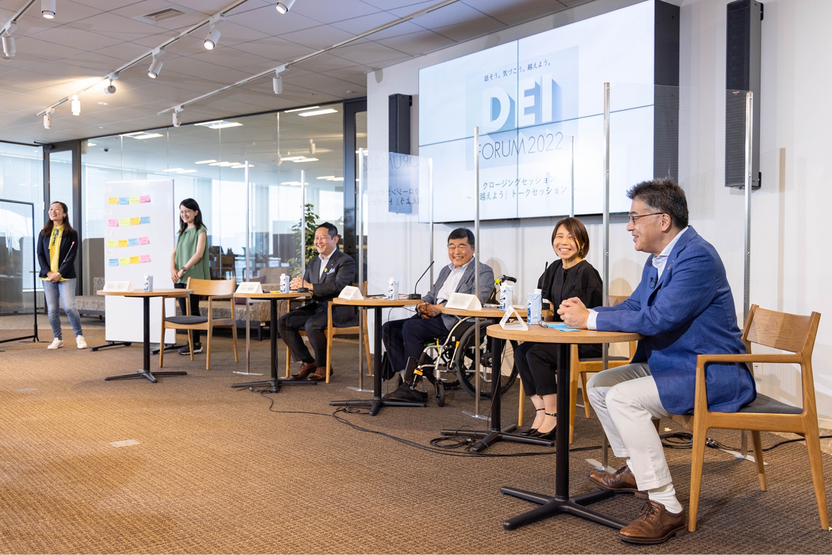 Photo: The talk session in progress. Two moderators are standing in the studio and five speakers are seated. They are chatting in a friendly atmosphere.