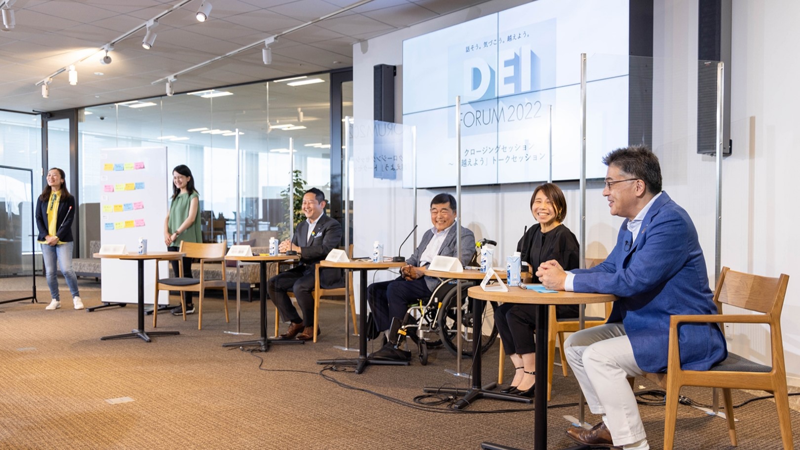 Photo: Group DEI Forum 2021. Moderator and speakers Yuki Kusumi and Shigeki Mishima. On the large screen in the background are several faces of employees participating online. The words "DEI FORUM" appear at the bottom of the photo.