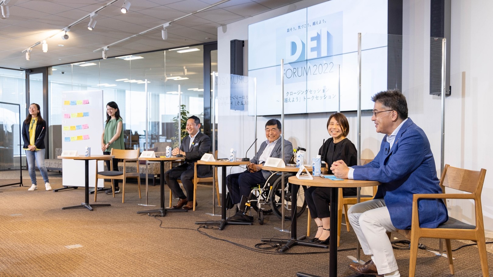 Photo: Group DEI Forum 2022 in progress. Two moderators are standing in the studio and five speakers are seated. They are chatting in a friendly atmosphere.