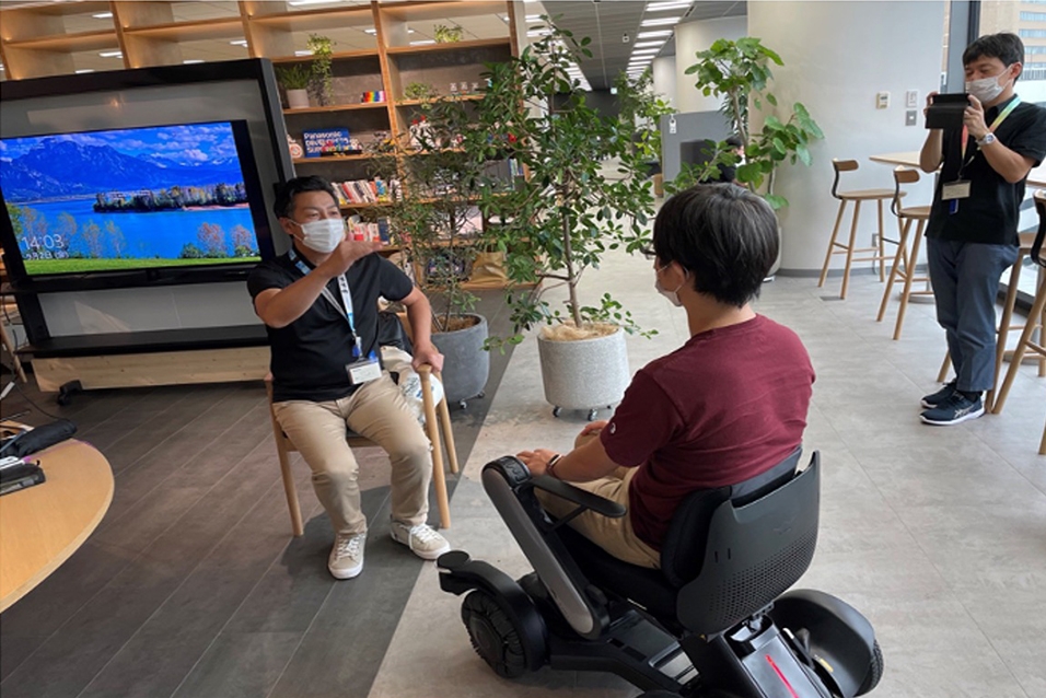 Photo: A participant sits on a chair at the same eye level as an electric wheelchair user and greets them.