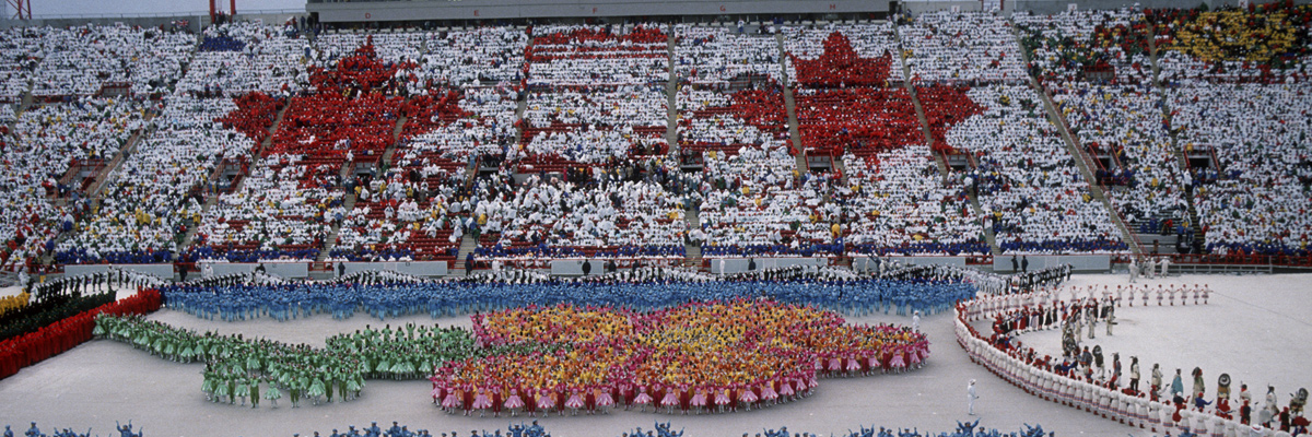照片：在卡尔加里冬季奥运会的开幕式上，主看台上由人组成的枫叶和穿着鲜艳服装的人们在体育场中央区域进行的舞蹈表演