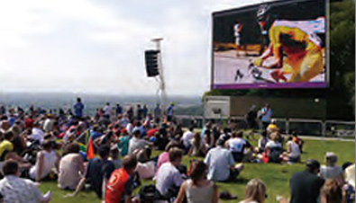 Photo: Spectators watching a road cycling competition shown on a large outdoor monitor